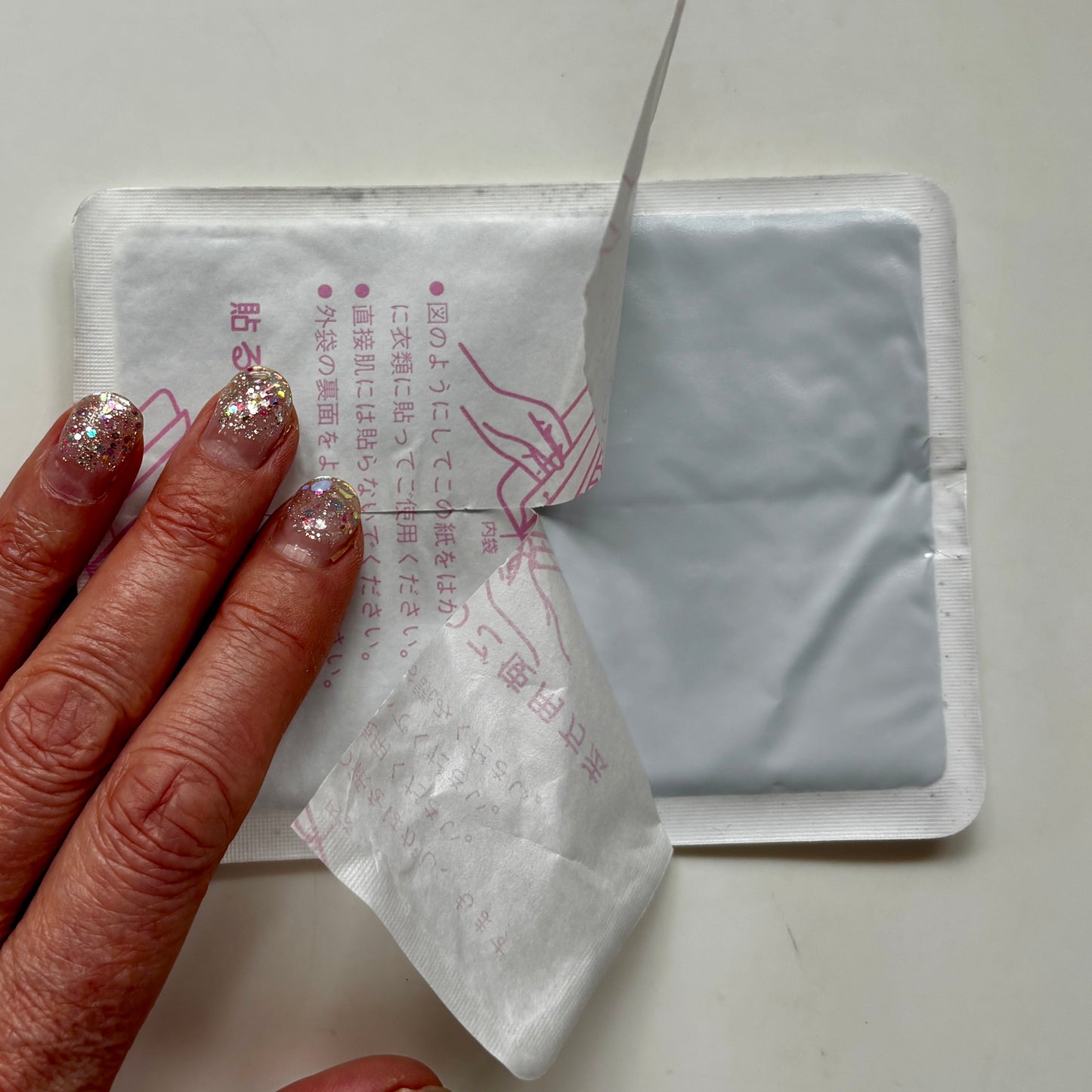 Close-up of a hand peeling back the paper backing of a Hotteeze Heat Pad to reveal the grey adhesive side, with Japanese instructions printed on the backing.